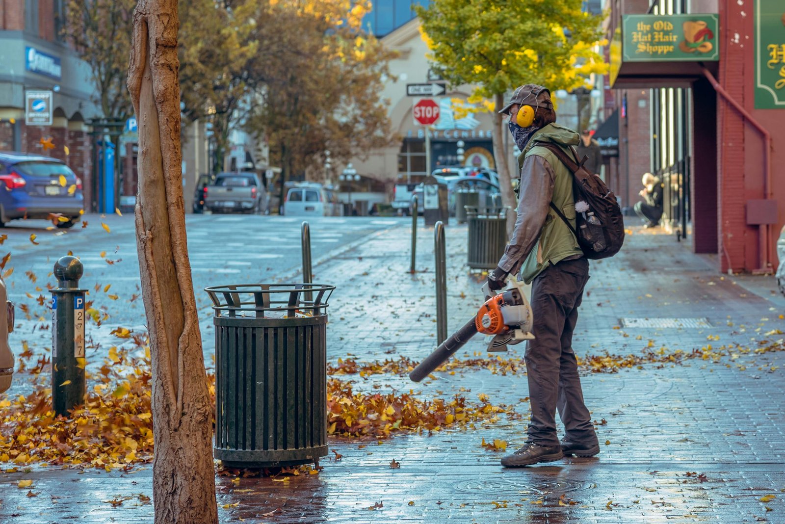 Street worker using a leaf blower during autumn in a city setting, surrounded by fallen leaves.