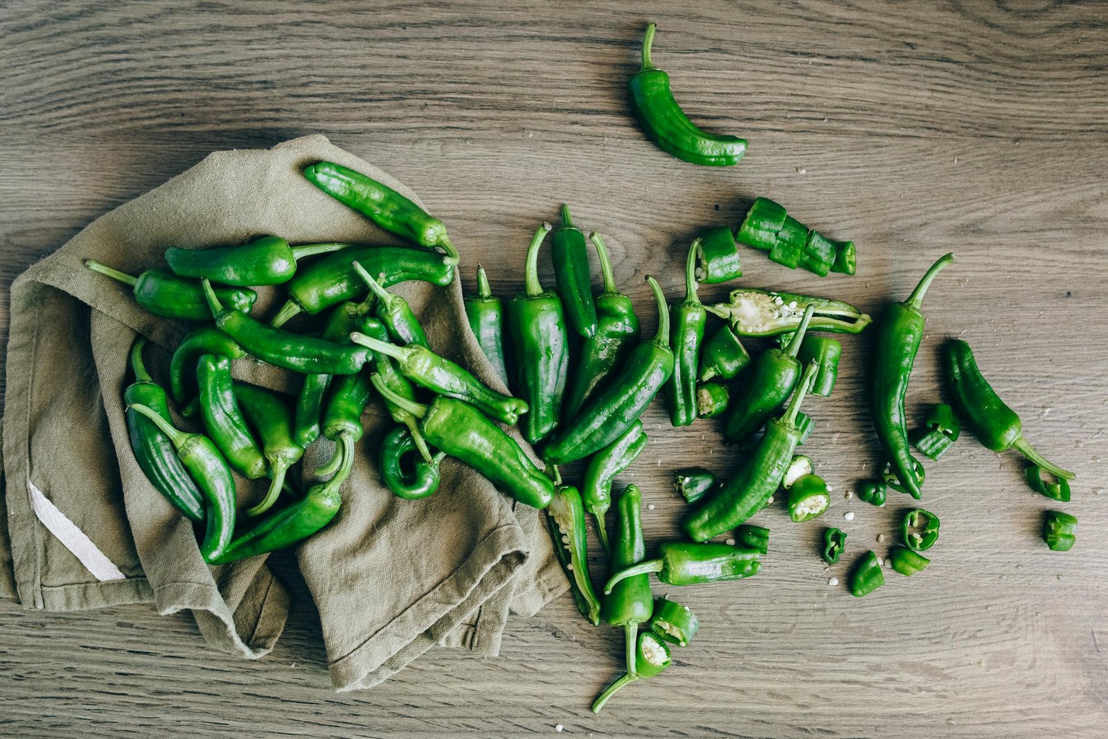 A flat lay of fresh green chilies on a wooden surface with a cloth.